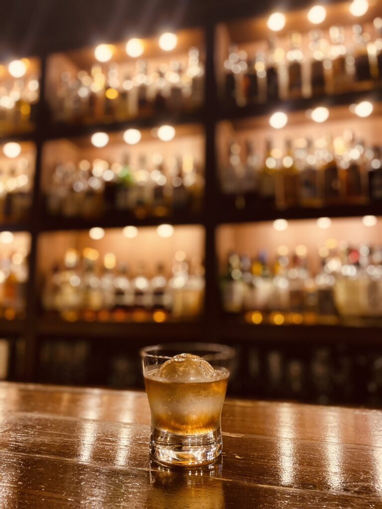 A relaxed evening at Bar Little Happiness in Hiroshima, featuring a sophisticated counter, warm lighting, and a massive shelf of 1,000+ whiskies.